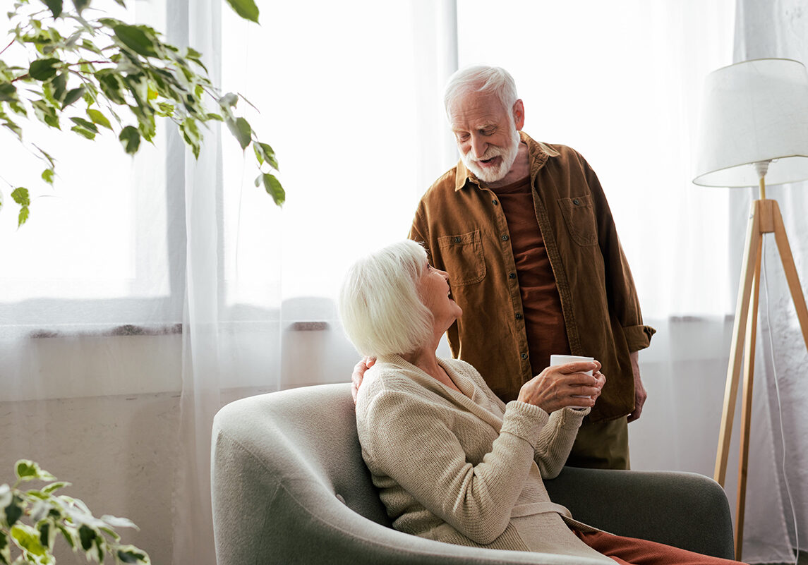 senior woman sitting in armchair with cup of tea while talking to smiling husband